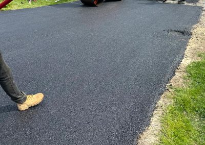 Workers using a steamroller to lay asphalt on a driveway next to a grassy lawn.