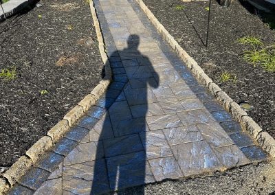 Shadow of a person on a curved, textured stone pathway with a walker nearby and garden on both sides.