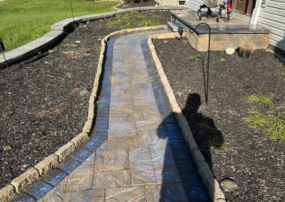 Stone pathway leads to a house entrance with a stroller on the porch, surrounded by grass and plants.
