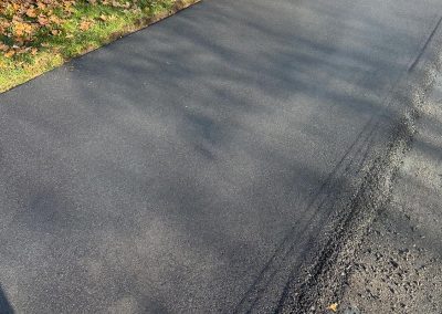 Freshly paved driveway next to a garden with shrubs and dry leaves on a sunny day.