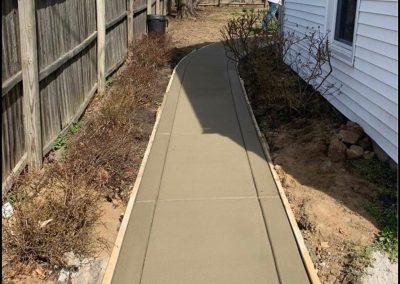 Freshly laid concrete path beside a white house, fenced on one side, with a person in the background.