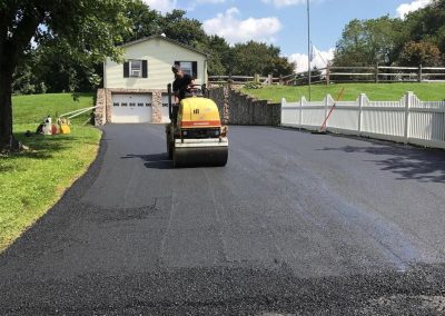 Person operating a roller compactor on a freshly paved driveway near a house, with greenery and fences around.