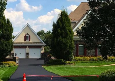Paved driveway with an orange cone and tape, leading to a two-car garage surrounded by trees and grass.