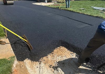 Workers paving a driveway with asphalt in front of a white house with red shutters.