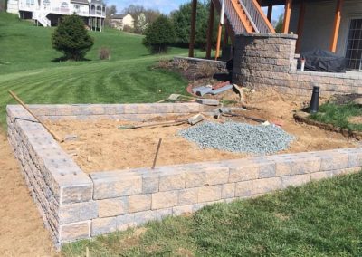 A backyard construction site with a partially built stone wall and a pile of gravel under a wooden deck.