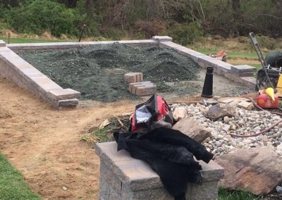 Garden area under construction with a stone border, soil, and tools nearby. Lawn and trees in the background.