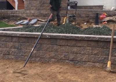 A person standing in a yard near landscaping tools and a pile of gravel, with a retaining wall in the foreground.