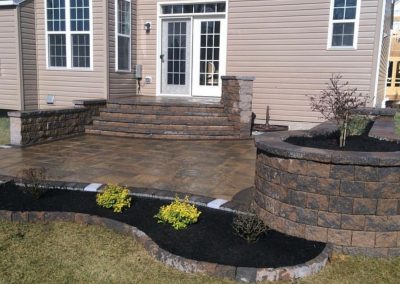 Backyard with a stone patio, raised garden beds, and steps leading to a house with beige siding and white-framed windows.