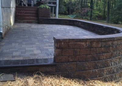 Brick retaining wall with a paved patio, surrounded by a forested area. Straw covers the ground in the foreground.