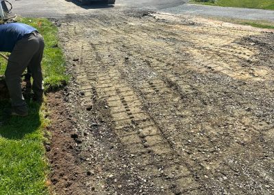 Construction site with earth-moving equipment clearing a driveway area, surrounded by houses and vehicles.