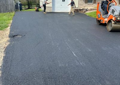 Workers paving a driveway with fresh asphalt in front of a two-story house. One operates a compactor machine.