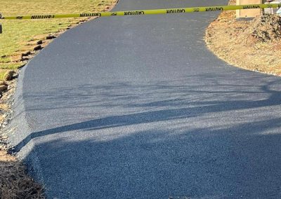 Freshly paved driveway leading to a garage, blocked by construction tape. Lawn and a small tree are visible.