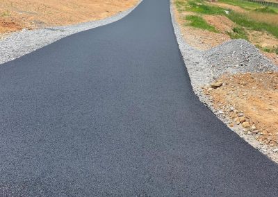 Freshly paved asphalt driveway leading to a gray house under construction with surrounding dirt mounds.