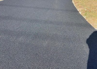 Freshly paved asphalt driveway curving through a yard with grass and a wooden fence.