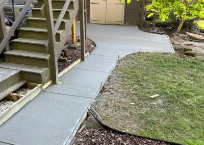 Freshly laid concrete path curves beside stairs and a shed, bordered by grass and a shrub.