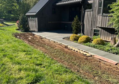Wooden house with black roof and a newly prepared dirt path surrounded by green grass and bushes.