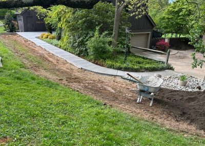 A freshly laid concrete pathway leads to a house surrounded by greenery, with a wheelbarrow nearby.
