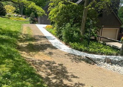 A sloped pathway leads to a house surrounded by green trees and grass on a sunny day.