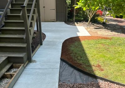 Concrete pathway beside brown stairs, leading to a wooden shed, with grass and brown pebbles along the sides.