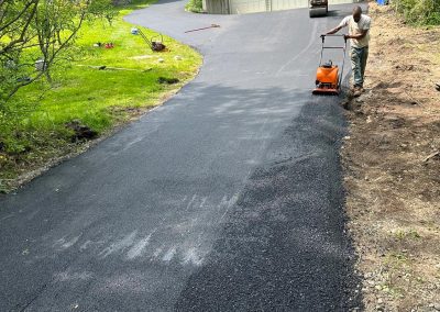 Person paving a driveway with a roller on a sunny day, surrounded by greenery and a gray, two-car garage.