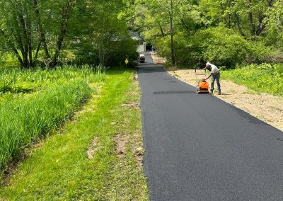 Person paving a rural path with an asphalt roller, surrounded by lush green trees and grass.