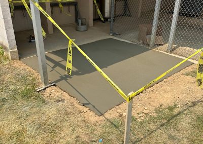 Freshly poured concrete slab surrounded by caution tape near a fenced area and building wall.