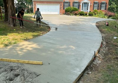 Two workers smooth wet concrete on a curved driveway in front of a large brick house surrounded by trees.