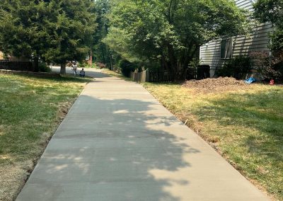 A freshly paved concrete path leading through a neighborhood lined with trees and a house on the right.