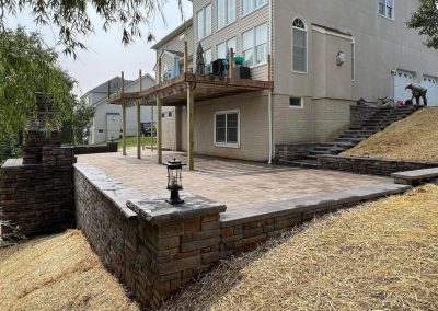 Backyard patio with stone retaining walls, a raised deck, and a lantern on a brick pillar near a multi-story house.