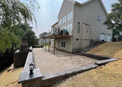 Spacious backyard patio with brick flooring, retaining walls, and a raised deck attached to a two-story house.