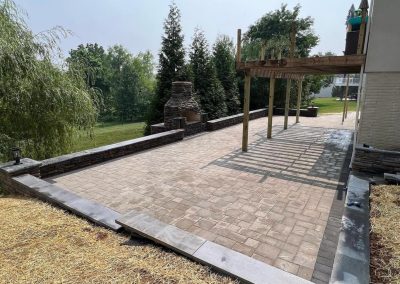 Paved patio with wooden pergola and brick fireplace, surrounded by trees and open lawn.
