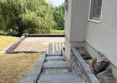 Stone stairs lead to a patio next to a house, surrounded by trees and grass, with bags of material on the side.