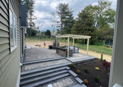 Backyard patio with stone tiles, pergola, brick barbecue, and steps leading to landscaped garden with trees.