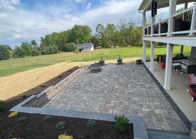 A backyard patio with a fire pit, seating area, and open grass field, surrounded by trees and a small shed.