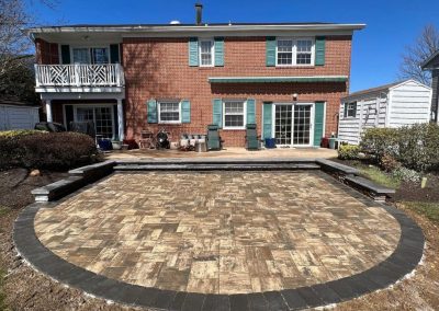 A brick house with a balcony overlooks a patterned stone patio under a clear blue sky.