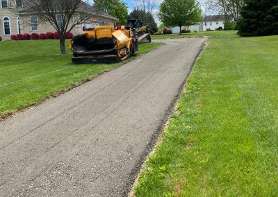 Paving machinery parked on a residential driveway with green grass and houses under a partly cloudy sky.