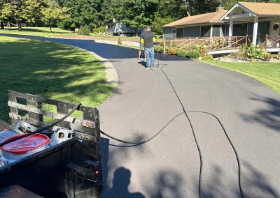 Person sealing a freshly paved driveway next to a house on a sunny day; trees in the background.