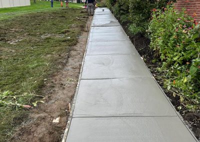 Freshly laid concrete sidewalk beside a red brick building, lined with bushes and grass. Workers visible in the distance.