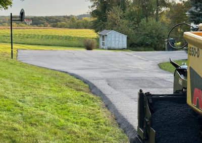 Paver on a road edge with fresh asphalt, surrounded by trees and open fields; small shed in the background.