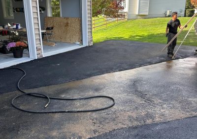 Person power-washing a driveway near an open garage with tools and plants, surrounded by a grassy yard.