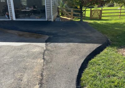Freshly paved driveway leading to an open garage, bordered by grass and a wooden fence.
