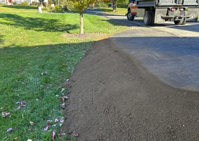 Freshly laid soil next to a driveway, with a dump truck and a tree with autumn leaves in the background.