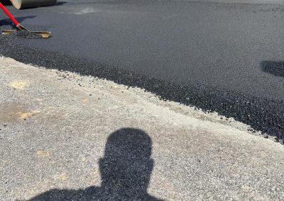 Shadow of a person on paved ground, with a steamroller smoothing fresh asphalt near a building.