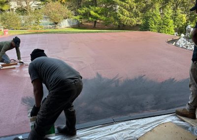 Workers applying black sealant on a large red rooftop surrounded by trees.