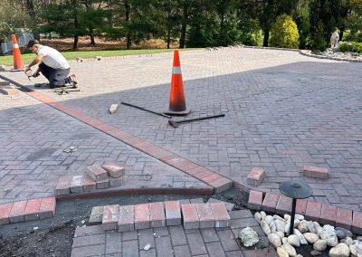 A person installing pavers on a driveway with orange cones and an edging of red bricks. Trees in the background.