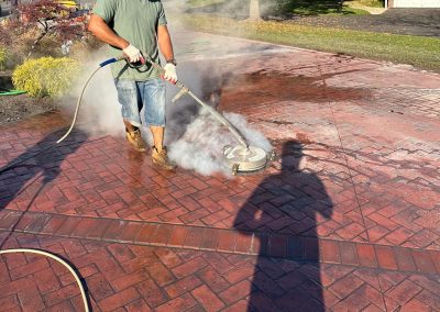 Person pressure washing a brick-patterned driveway, surrounded by steam, with trees and houses in the background.