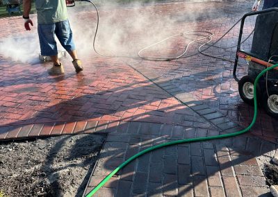 Two people power-washing a red brick patio, with steam rising, surrounded by trees and a garden hose.