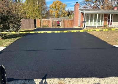 Freshly paved driveway with caution tape, surrounded by autumn trees and a house in the background.