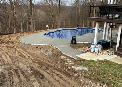 Workers installing concrete around a backyard pool, with trees and a house in the background.