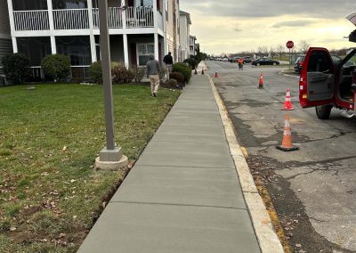 Freshly paved sidewalk beside a residential building, traffic cones and an open car trunk are visible. Cloudy sky above.